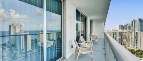 Un balcón con amplias ventanas que ofrecen vistas a la ciudad, una pequeña mesa y dos sillas de mimbre.