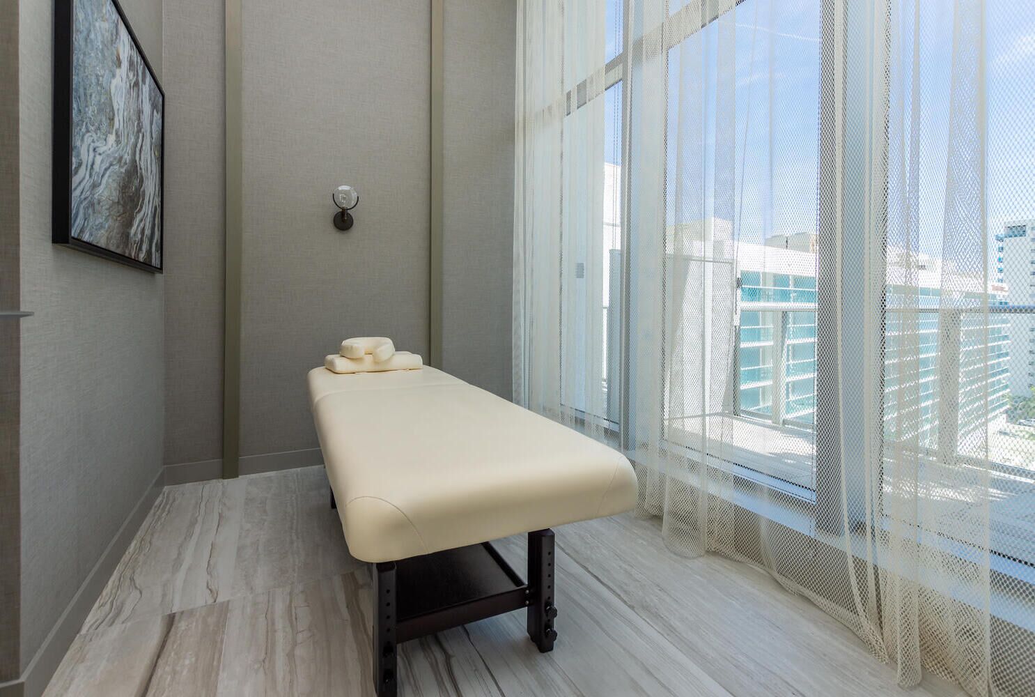 A room with a massage table, a wall-mounted massage device, and large windows with sheer curtains.