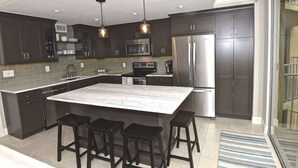 A modern kitchen with dark cabinets, a large island with stools, stainless steel appliances, and pendant lighting. - Renovated  Condo with Luxury Amenities in Perfect Beach Location (Marco Island)