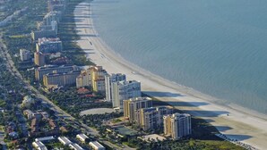 An aerial view of a coastal area with high-rise buildings along the beachfront, a sandy beach, and a calm sea. - Renovated  Condo with Luxury Amenities in Perfect Beach Location (Marco Island)