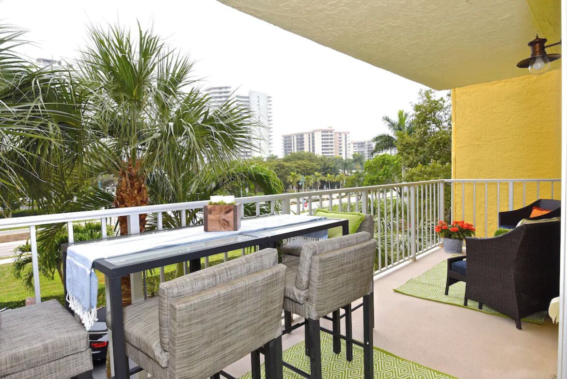 Een balkon met een tafel en stoelen, een potplant en uitzicht op palmbomen en gebouwen.