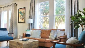Ein Wohnzimmer mit einem braunen Ledersofa, einem blauen Sessel, einem Holztisch und einem großen Fenster mit Vorhängen.