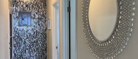 Un couloir avec un mur orné de motifs, un miroir rond et une porte donnant sur une autre pièce.
