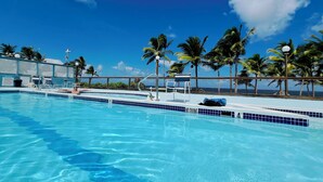 Piscina azul límpida com placa de 'NÃO MERGULHAR', rodeada por palmeiras e vista para o oceano.