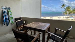 Uma varanda com mesa e cadeiras de madeira, um letreiro com a palavra 'Welcome' e vista para uma praia com palmeiras.