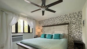 Quarto com cama, ventoinha no teto, janela com cortinas, mesa de cabeceira e parede com padrão em mosaico.