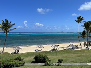 Una playa con agua turquesa cristalina, palmeras y una zona verde con sillas de descanso y sombrillas.