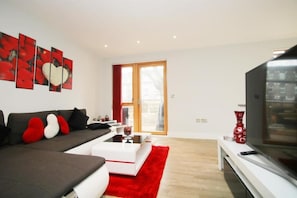 Relax in this stylish living room with a large flat-screen TV, comfortable sofa, and glass door leading to an outdoor area.