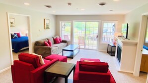 Ein Wohnzimmer mit roten Sofas, einem Couchtisch und einem Fernseher. Es gibt einen Balkon mit Blick ins Freie.