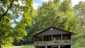 Een rustieke hut met een veranda, omgeven door weelderige begroeiing en bomen.