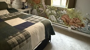 Een slaapkamer met een groot bed, een nachtkastje, een lamp, een raam met gordijnen en een muurschildering van een boslandschap.