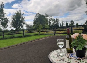 Une table avec une bouteille de vin et un verre, une plante en pot, et une vue sur un paysage rural avec une clôture et des arbres.