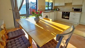 Un coin repas avec une table en bois et des chaises rembourrées, une cuisine en arrière-plan et une vue sur l’extérieur.