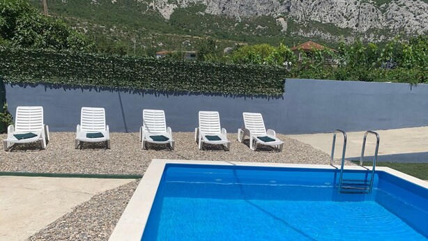 A rectangular swimming pool with a clear blue water surface, surrounded by white lounge chairs with green cushions. - Natura-Gata,Omiš-small stone apartman with swimming pool (Gata)