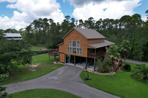 A two-story wooden house with a balcony, surrounded by a well-maintained lawn and trees. - Pool, Boat Ramp, Games, Pass Christian Waterfront Sleeps 18 Guests 6 Bedrooms  (Pass Christian)