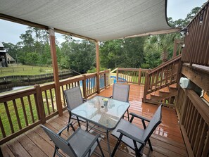 A covered outdoor deck with a glass table and chairs, wooden railings, and a view of a swimming pool and greenery. - Pool, Boat Ramp, Games, Pass Christian Waterfront Sleeps 18 Guests 6 Bedrooms  (Pass Christian)