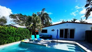 Ein Swimmingpool mit zwei blauen Liegestühlen, einer überdachten Sitzgelegenheit und einem Gebäude mit rotem Dach.