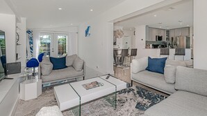 Ein modernes Wohnzimmer mit zwei grauen Sofas, einem weißen Couchtisch und einem Couchtisch mit Glastischplatte. Durch einen offenen Durchgang ist eine Küchenzeile erkennbar.