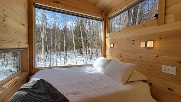 Una habitación de cabaña de madera con una cama, ventanas amplias y vistas a un bosque nevado.
