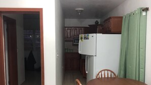 A dining area with a wooden table and chairs, a kitchen with wooden cabinets and a refrigerator, and a hallway leading to another room.