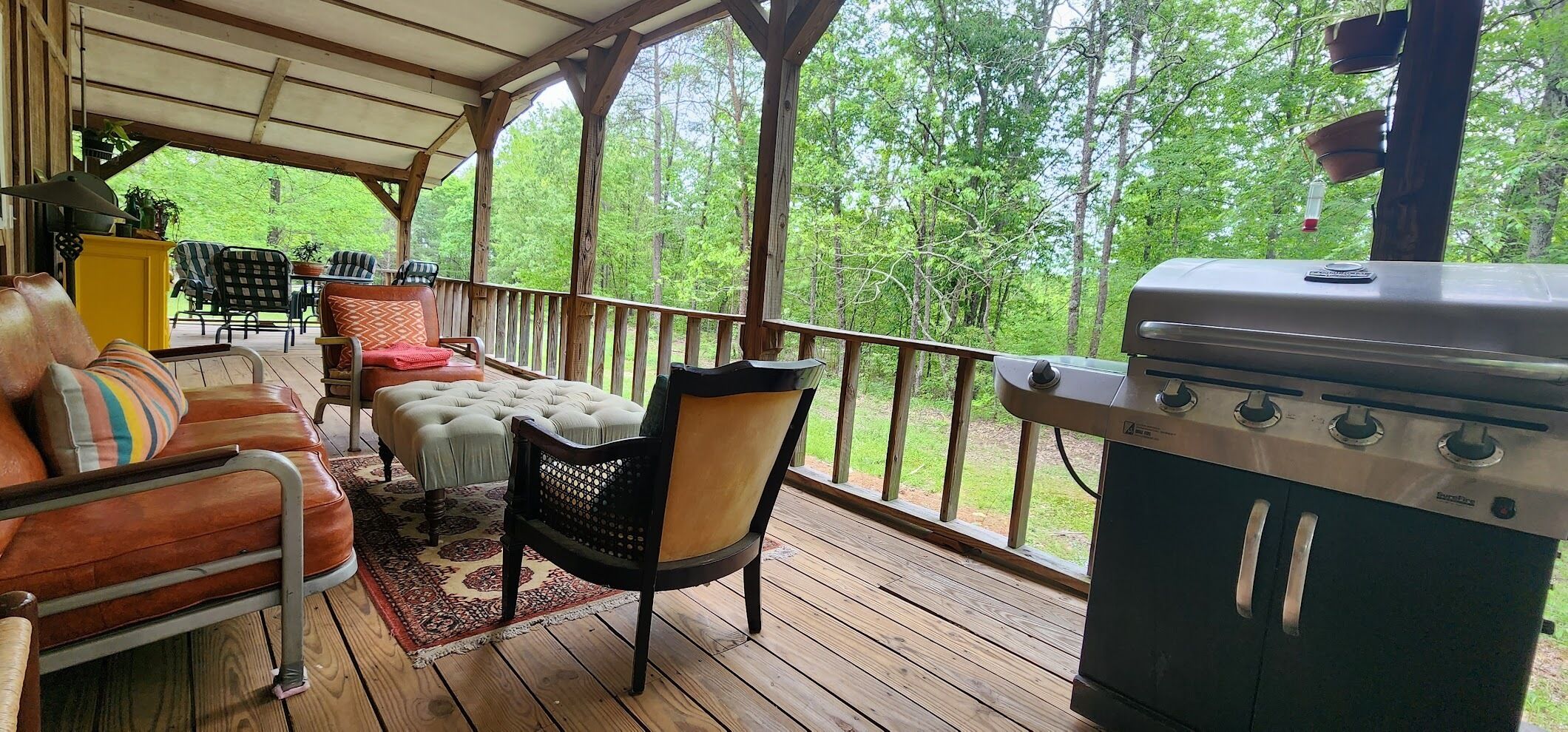 A covered outdoor deck with a grill, seating area with a sofa and chairs, and a view of a forested area.