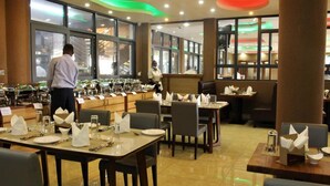 A restaurant with multiple dining tables, some with white napkins and cutlery, and a person in a white shirt standing near the entrance. - Hera Hotel (Kampala)