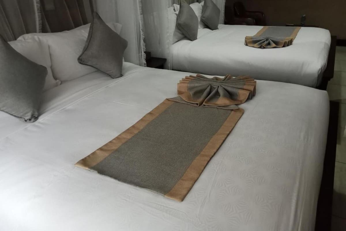 A hotel room with two beds, each with white linens and grey pillows. A brown and grey throw is placed on the bed in the foreground.