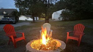 Ein rundes Feuerbecken mit zwei roten Adirondack-Stühlen davor, umgeben von einer Rasenfläche mit Bäumen und einem Gebäude im Hintergrund.