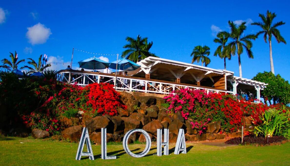 Un bâtiment sur lequel on peut lire l’inscription « ALOHA », situé devant un paysage rocheux, avec des fleurs rouges et des palmiers en arrière-plan.