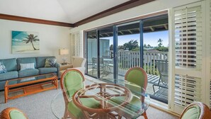 A living room with a sofa, coffee table, and chairs, a painting of a palm tree, and a balcony with a view of the outdoors. - Luxury Upgraded Condo #98, AC, Poipu Beach, Partial Ocean View (Koloa)