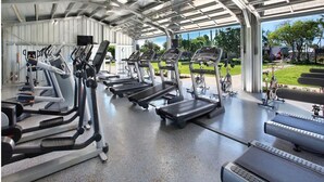 A covered gym area with multiple treadmills and exercise bikes, a concrete floor, and a view of a park and buildings outside. - Luxury Upgraded Condo #98, AC, Poipu Beach, Partial Ocean View (Koloa)