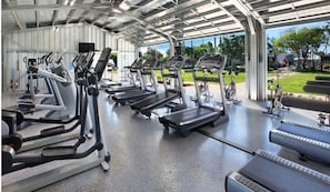 A covered gym area with multiple treadmills and exercise bikes, a concrete floor, and a view of a park and buildings outside. - Luxury Upgraded Condo #98, AC, Poipu Beach, Partial Ocean View (Koloa)