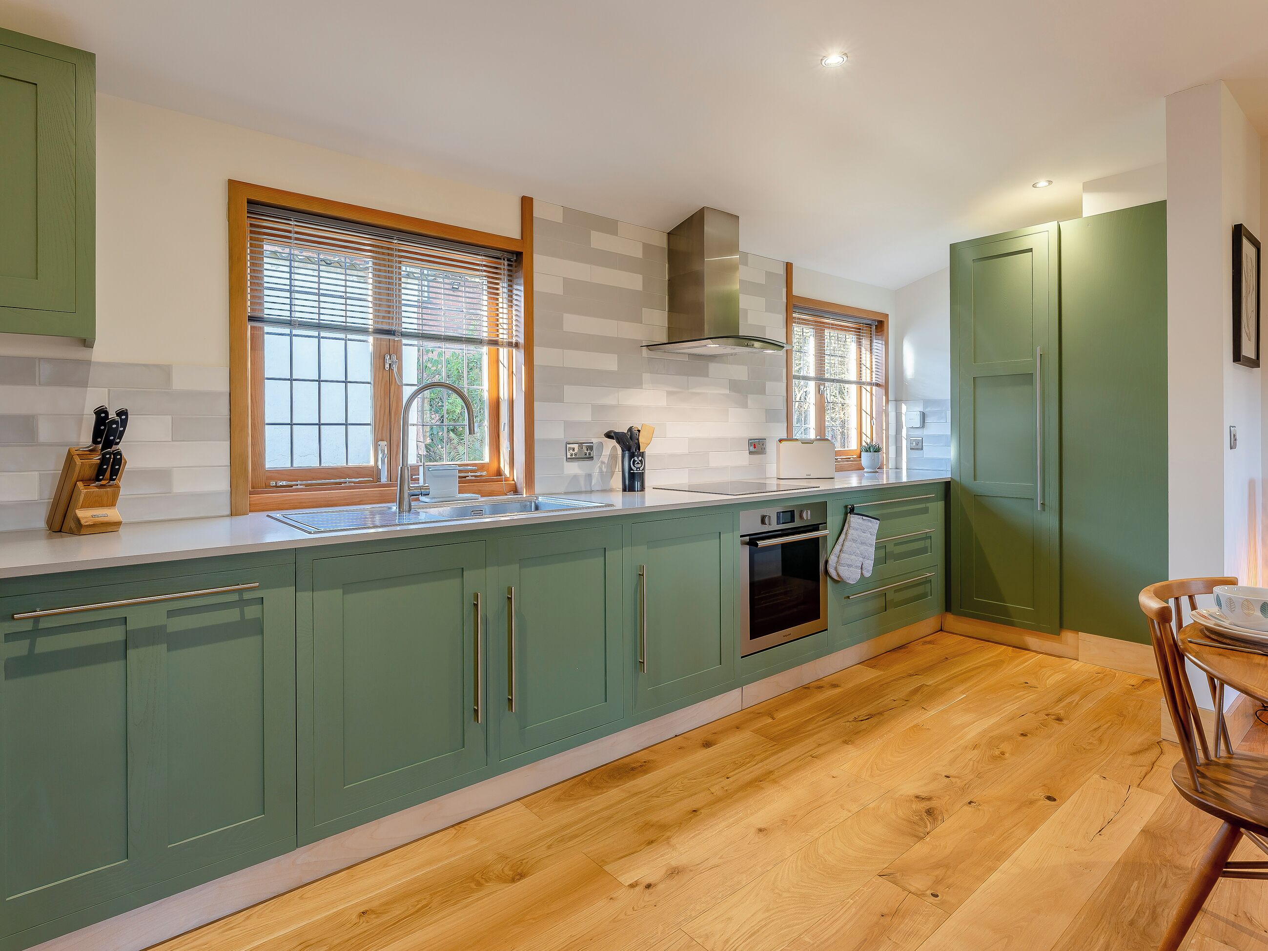 Une cuisine avec des meubles de couleur verte, un plancher en bois, un four encastré et une fenêtre avec des stores.
