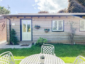 A wooden house with a front door, two windows, and a small garden with a table and chairs. - Bluebell Lodge-UK32958 (Ashford)