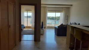 A spacious living area with a large sliding glass door leading to a balcony, a dark sofa, a small white table with a TV, and a bar area with stools. - AIRPORT to Comme à la maison 2, 5 minutes from the airport (Nouaceur)