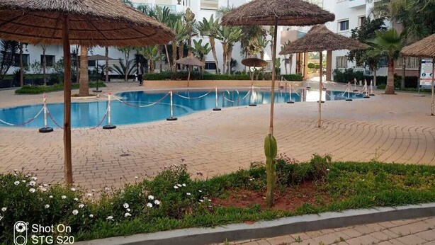 A swimming pool area with thatched umbrellas, palm trees, and a brick pathway. - AIRPORT to Comme à la maison 2, 5 minutes from the airport (Nouaceur)
