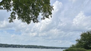 Une terrasse au bord du lac, avec un foyer intégré et des chaises Adirondack rouges, donnant sur un lac où l’on aperçoit un bateau et une rive éloignée.