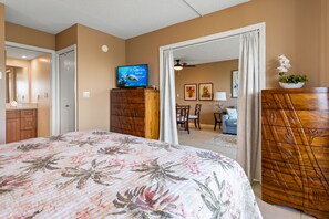 A bedroom with a floral bedspread, a television on a wooden cabinet, a dining area with chairs and a table, and a view into another room with a couch and a plant.