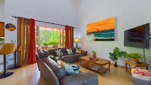 A modern living room with a sofa, coffee table, and TV. There is a painting on the wall and a plant on a side table. - Casa Tropical, Spacious 3 BR Home with Pool (Brasilito)