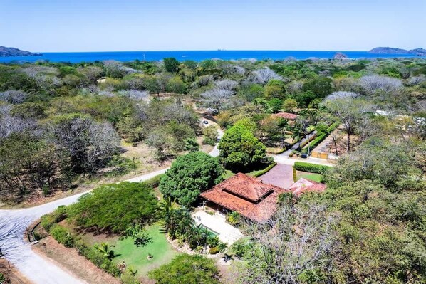 A large, open landscape with a central building surrounded by dense vegetation and a clear view of the ocean.