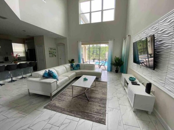 A spacious living room with a white sectional sofa, a coffee table, and a TV on a white console. There is a large window providing natural light and a view of the outdoors. - Villa, private pool, water park, Orlando parks (Kissimmee)