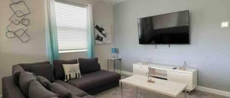 Una sala de estar moderna con un sofá seccional, un televisor de pantalla plana montado en la pared, un ventilador de techo y una ventana con persianas.