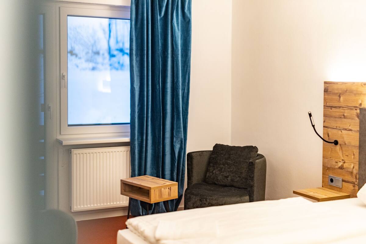 Une chambre à coucher comprenant un lit, une chaise, une fenêtre avec des rideaux, un radiateur et un panneau mural en bois.