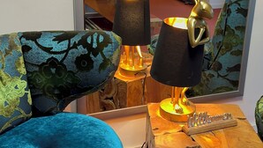 Un salon chaleureux avec un fauteuil en velours bleu, une table basse en bois surmontée d’une lampe, et un miroir qui reflète la pièce.