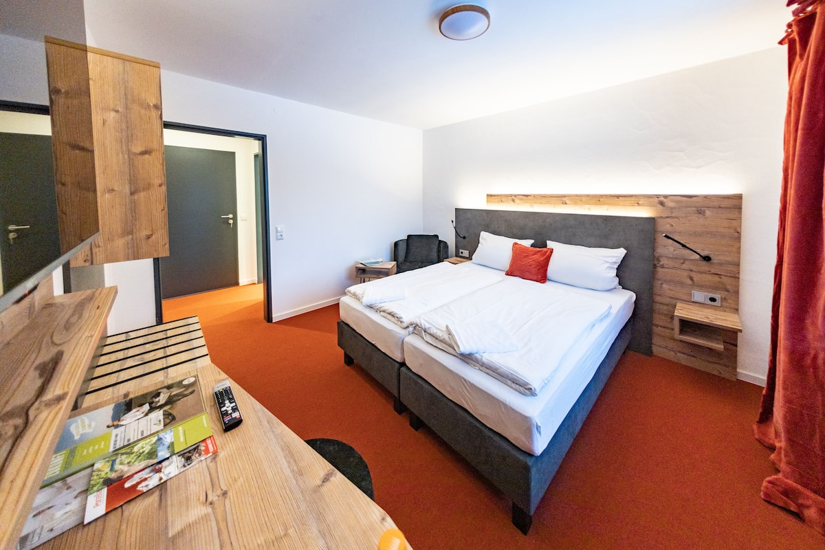 Une chambre d’hôtel moderne avec un grand lit, un bureau avec des livres, une chaise et une tête de lit en bois.