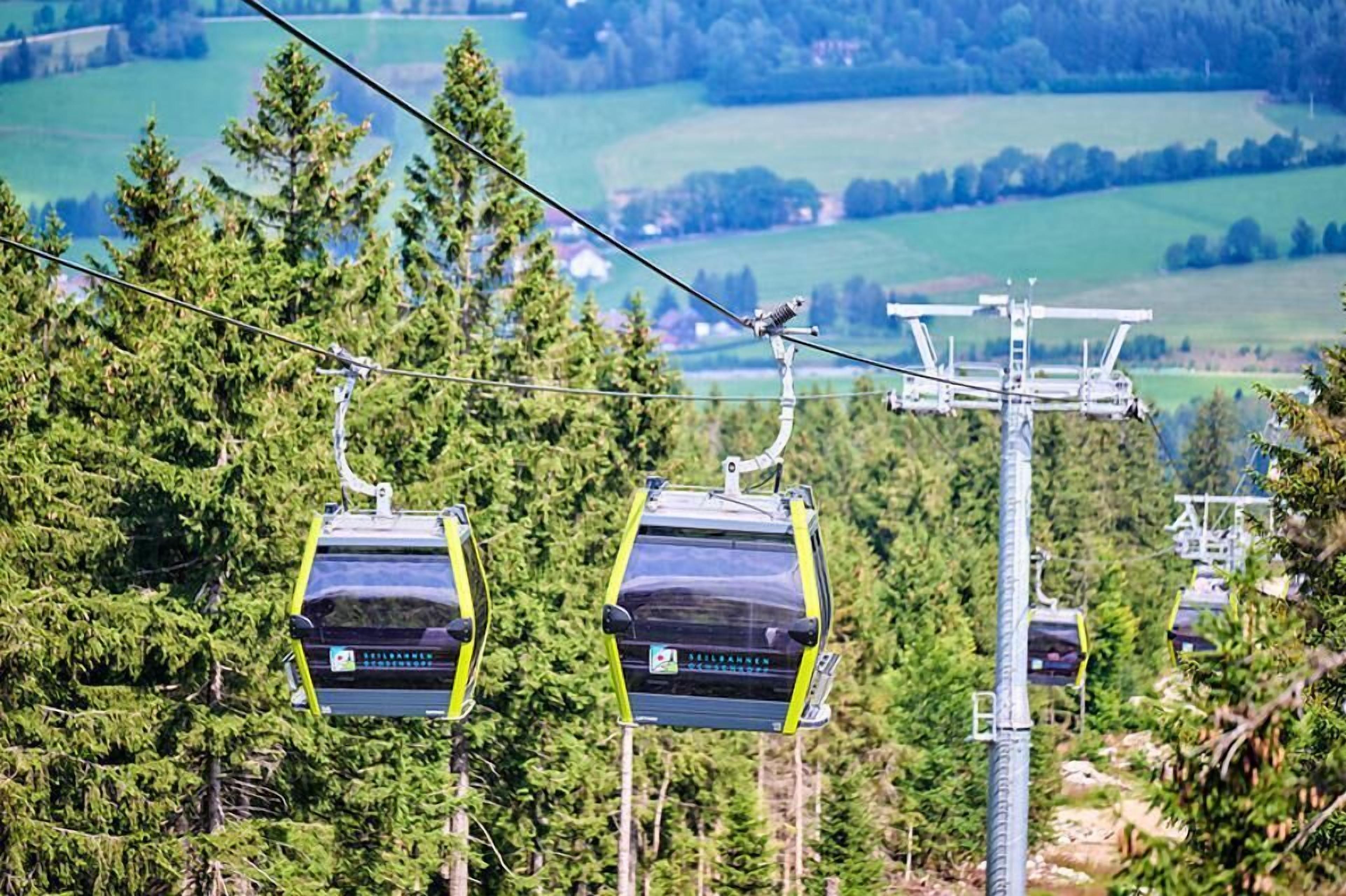 A cable car system with cabins suspended by cables, set against a backdrop of green fields and trees.