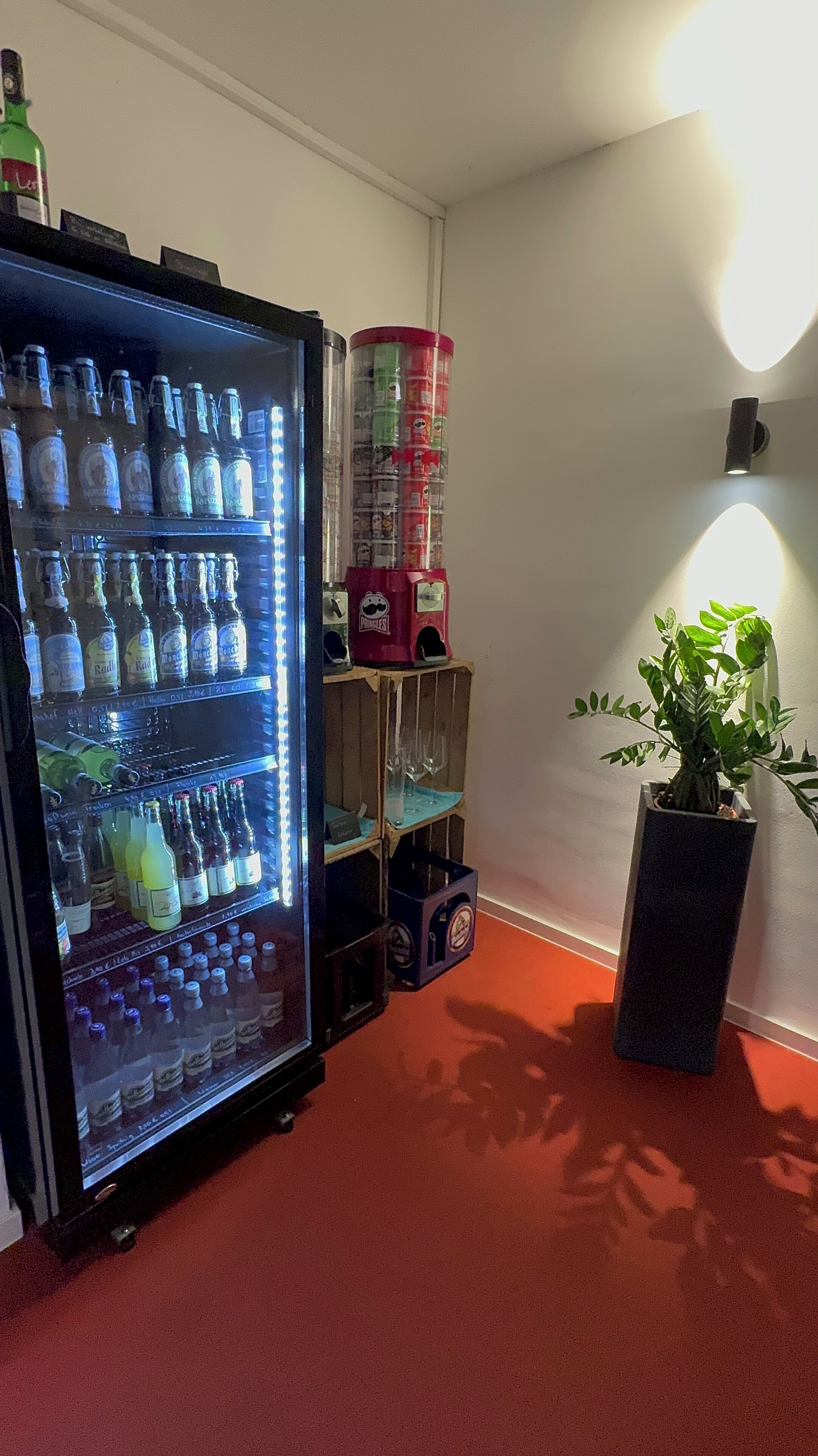 A room with a minibar stocked with various beverages, a stack of Pringles cans, and a potted plant.