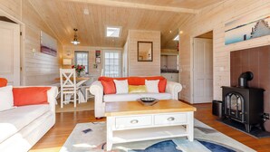 A cozy cabin interior with a wooden ceiling, a dining area, a living room with a sofa and coffee table, and a wood-burning stove.