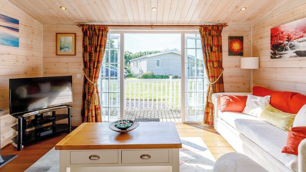 A cozy living room with a wooden ceiling, a television, a coffee table with a bowl, a sofa with colorful cushions, and a large window with curtains.