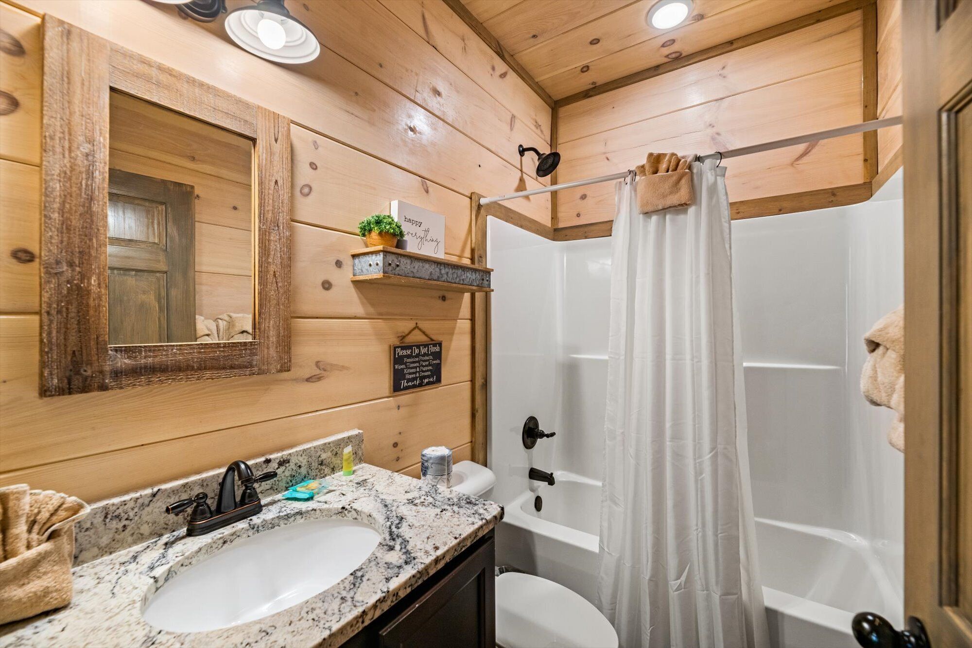 Casa de banho de cabana de madeira com lavatório de bancada em granito, espelho, prateleira com artigos de higiene pessoal, cortina de chuveiro e sanita.
